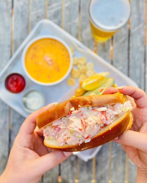 A person holds a lobster roll with a bowl of soup and a beer in the background, set on a wooden table.