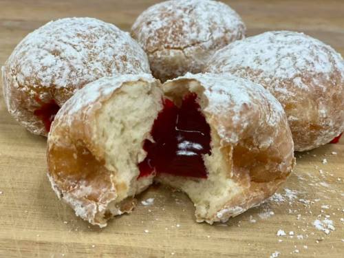 A close-up of powdered sugar doughnuts, one cut open to reveal a red jelly filling inside.