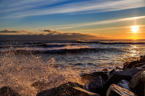 Waves crash against rocky shore at sunset, with vibrant colors in the sky reflecting on the water.