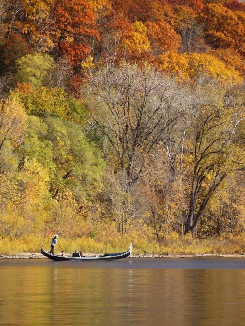 A canoe with two people paddling on a calm river, surrounded by vibrant autumn foliage.