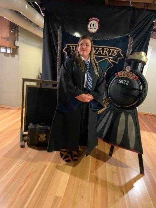 A person in a Hogwarts robe and tie stands beside a train display, with a "Platform 9¾" banner in the background.