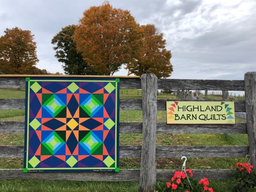 A colorful quilt pattern displayed on a wooden fence, with autumn trees and a sign reading "Highland Barn Quilts."