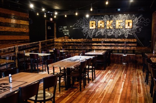 Interior of a cozy restaurant named "BAKED," featuring wooden tables, benches, and a decorative wall with lights.