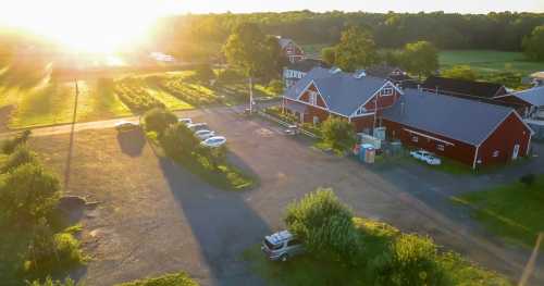 Aerial view of a farm at sunset, featuring red barns, green fields, and parked cars along a gravel driveway.