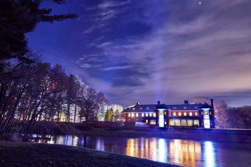 A large, illuminated building by a calm lake at night, surrounded by trees and a cloudy sky.