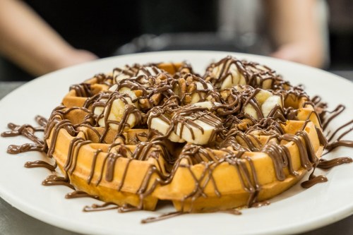 A delicious waffle topped with banana slices and drizzled with chocolate sauce, served on a white plate.