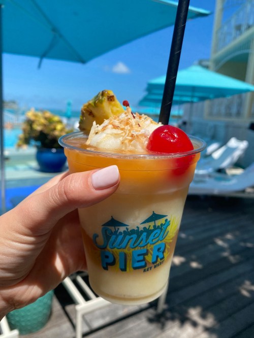 A hand holds a tropical drink with a cherry and pineapple, against a sunny poolside backdrop with blue umbrellas.
