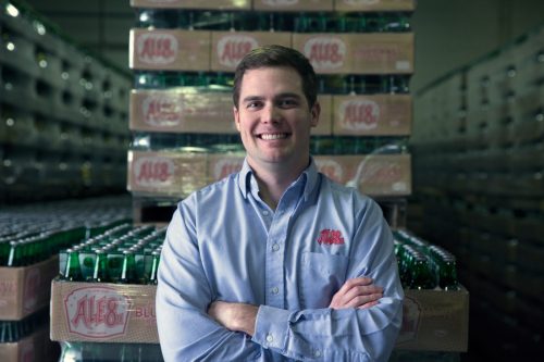 A smiling man in a blue shirt stands confidently in a warehouse filled with stacked beverage crates.