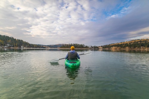 A person in a green kayak paddles on a calm lake surrounded by trees and mountains under a cloudy sky.