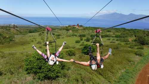 Two people ziplining side by side, holding hands, over a lush green landscape with mountains and ocean in the background.