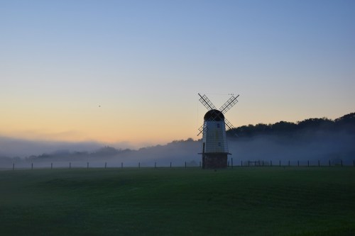 A windmill stands in a misty field at dawn, with soft colors of sunrise in the background.
