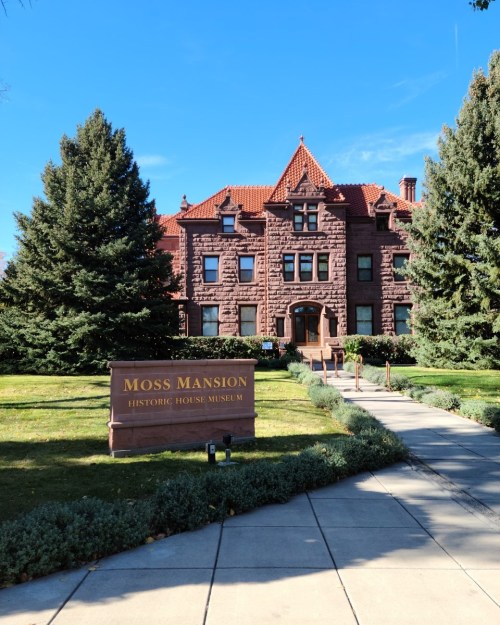Historic Moss Mansion with red stone architecture, surrounded by trees and a well-maintained lawn. Clear blue sky above.