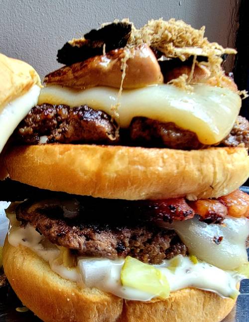 A towering burger with multiple beef patties, melted cheese, bacon, and toppings, served between two toasted buns.