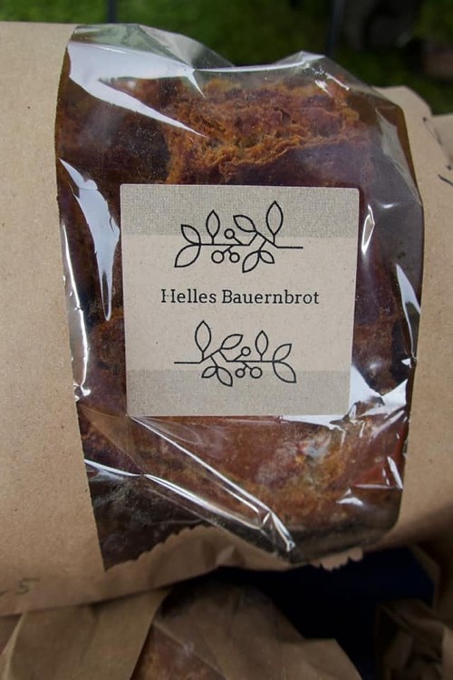 A loaf of bread wrapped in clear plastic with a label reading "Helles Bauernbrot" and decorative leaf designs.