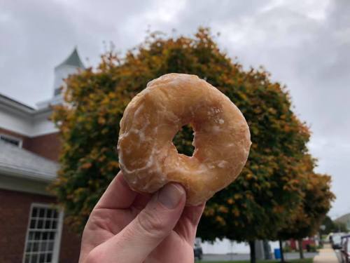 A hand holds a glazed donut in front of a tree with autumn leaves and a building in the background.