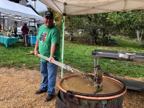 A man in a green shirt stirs a large pot outdoors, with tents and tables in the background.