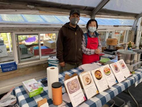 A man and woman wearing masks stand behind a table with food menus in a greenhouse setting.