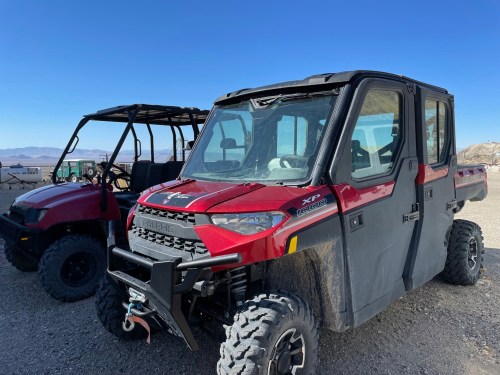 Two off-road vehicles parked on rocky terrain under a clear blue sky. One is a red enclosed model, the other is a red open model.