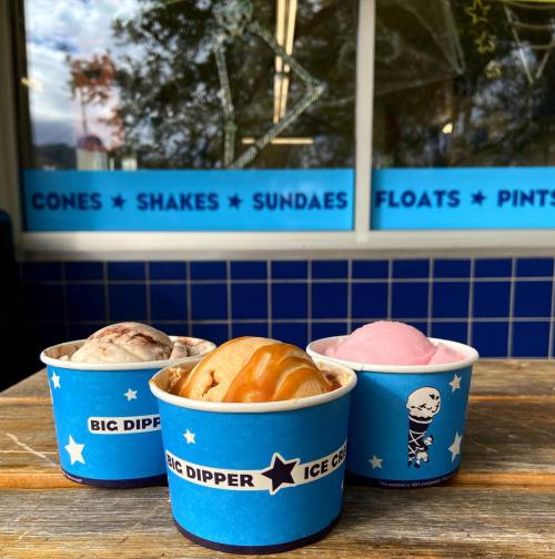 Three colorful ice cream cups on a wooden table outside an ice cream shop, featuring various flavors.