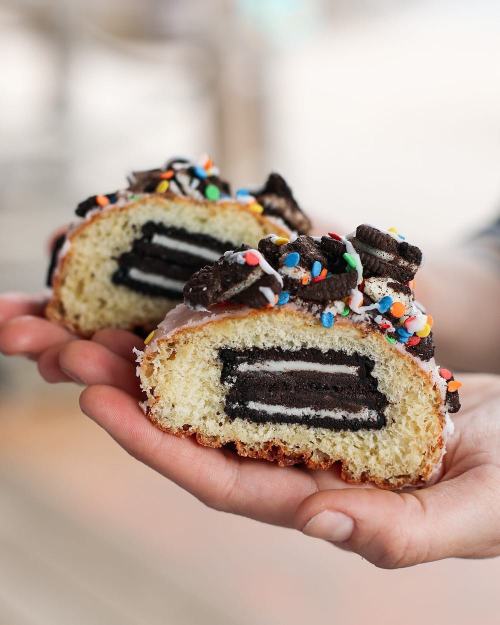 A hand holding a sliced dessert revealing layers of cake, Oreo cookies, and colorful sprinkles on top.