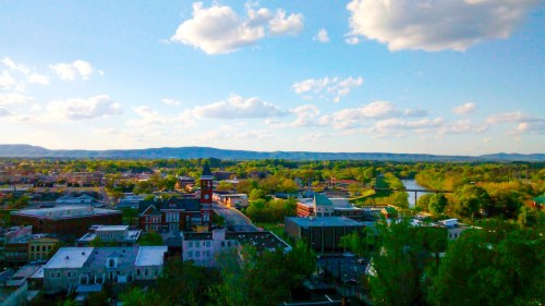 A scenic view of a town with green trees, a river, and a clock tower under a blue sky with fluffy clouds.