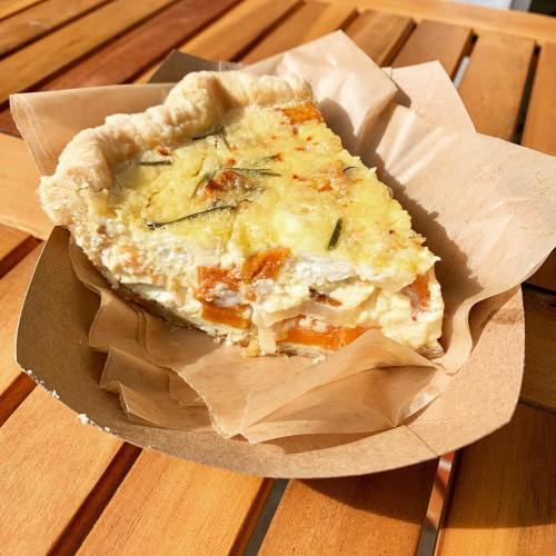 A slice of quiche in a paper dish, featuring a golden crust and a filling with vegetables and herbs.