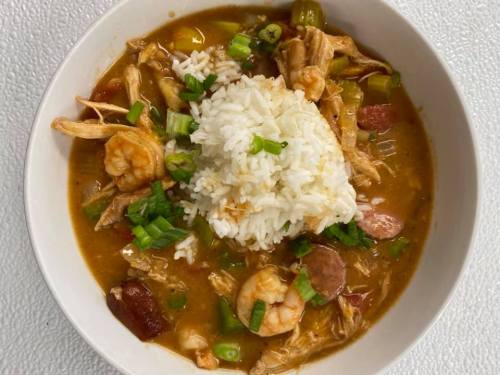 A bowl of gumbo with shrimp, chicken, sausage, and green onions, topped with a mound of white rice.