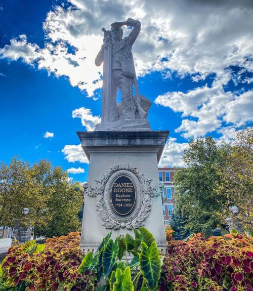 Statue of Daniel Boone, an explorer, surrounded by colorful plants and a bright blue sky with clouds.