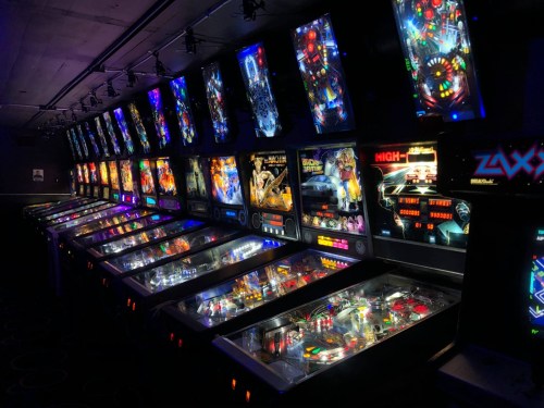 A row of colorful pinball machines illuminated in a dark arcade, showcasing various game designs and lights.