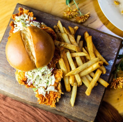 A crispy fried chicken sandwich with coleslaw on a wooden board, accompanied by golden French fries.