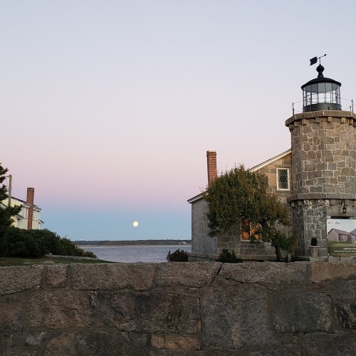 A stone lighthouse beside a calm sea at dusk, with a rising moon and pastel sky in the background.
