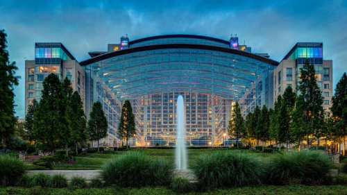 A modern glass building with a large arch, surrounded by trees and a fountain, illuminated at dusk.