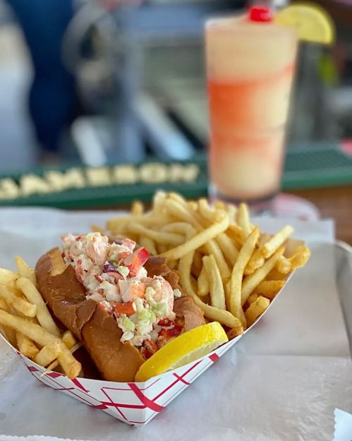 A lobster roll with fries and a colorful drink in the background, served on a tray.