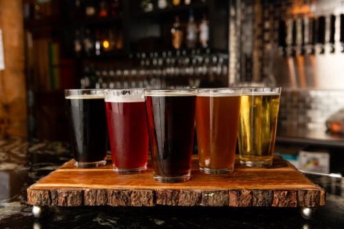 A wooden tray holds six glasses of different beers, showcasing a variety of colors and styles.