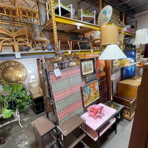 A cluttered thrift store display featuring various furniture items, lamps, and decorative pieces on shelves.