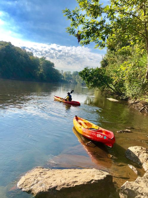 A person kayaking on a calm river, surrounded by trees and rocky banks under a blue sky.