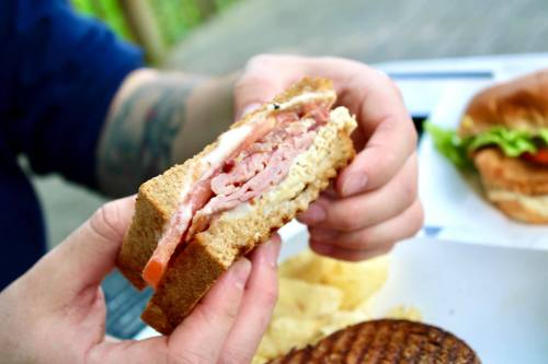 A person holds a sandwich filled with layers of meat and vegetables, with chips and another sandwich in the background.