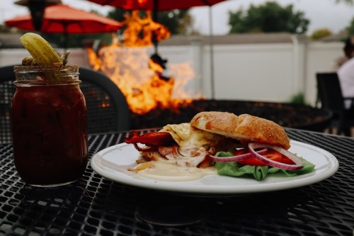 A sandwich with bacon, cheese, and veggies on a plate, next to a cocktail in a jar, with a fire pit in the background.