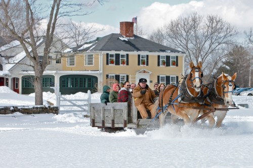 A horse-drawn sleigh carries passengers through snowy terrain, with a charming house in the background.