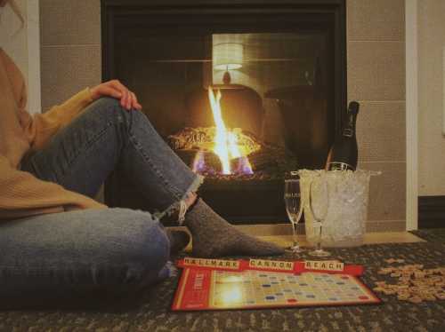 Cozy scene by a fireplace with a Scrabble game, champagne, and a person in casual attire enjoying a relaxing evening.