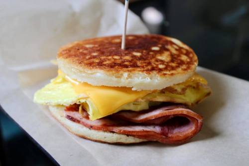 A sandwich with a fluffy pancake, cheese, egg, and ham, held together with a toothpick on a paper wrapper.