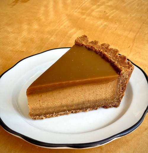 A slice of creamy caramel cheesecake on a graham cracker crust, served on a white plate with a black rim.