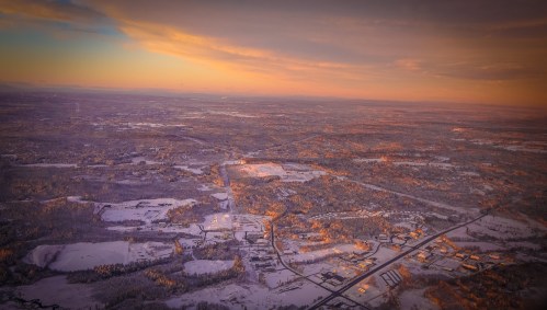 Aerial view of a snowy landscape at sunset, with fields, roads, and distant mountains under a colorful sky.