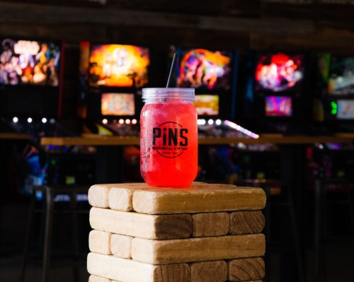 A colorful drink in a mason jar sits atop a wooden block, with pinball machines blurred in the background.