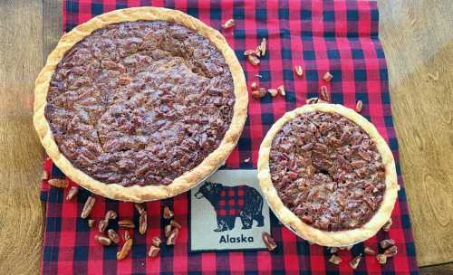 Two pecan pies, one large and one small, on a red and black checkered cloth with scattered pecans and an Alaska-themed napkin.