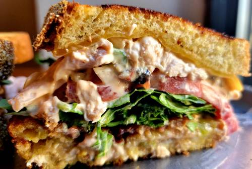 Close-up of a layered sandwich with greens, meats, and creamy sauce, showcasing its hearty ingredients.