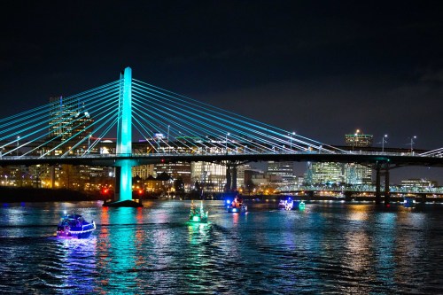 A brightly lit bridge at night with colorful boats on the water, reflecting city lights in the background.