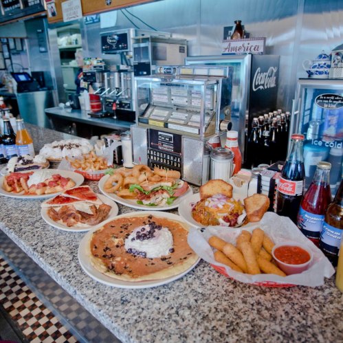 A diner counter filled with various dishes, including pancakes, fried foods, and drinks, showcasing a classic American menu.