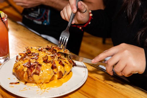 A person cuts into a loaded baked potato topped with cheese and bacon, with a drink on the side.