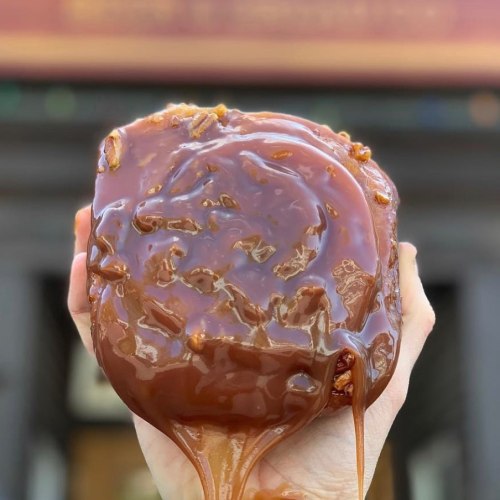 A close-up of a hand holding a sticky, caramel-coated dessert with a glossy finish.